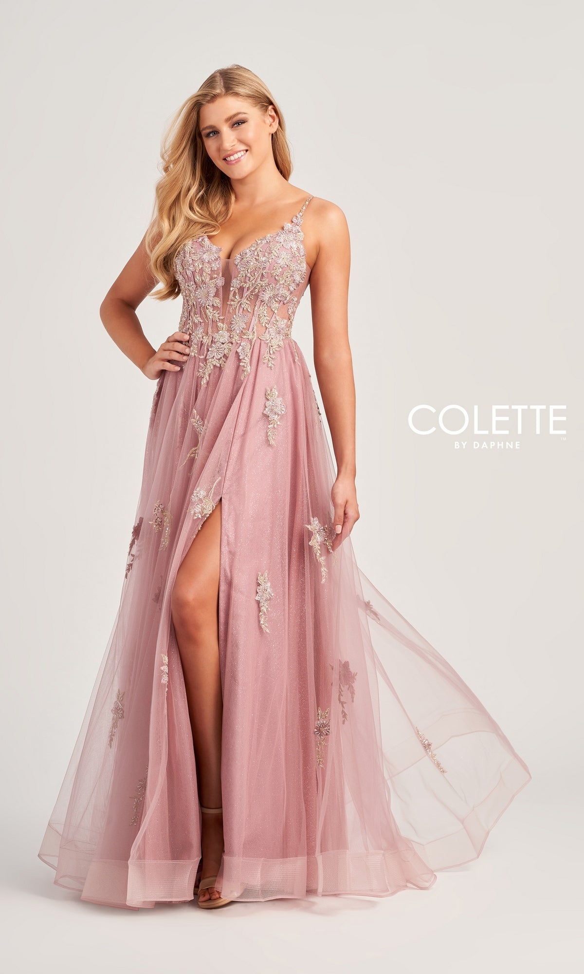 A woman stands and smiles against a plain background, wearing the Glitter-Lace Colette Designer Prom Gown CL5143—a mauve, floor-length evening dress with floral lace, embellishments, and a thigh-high slit.